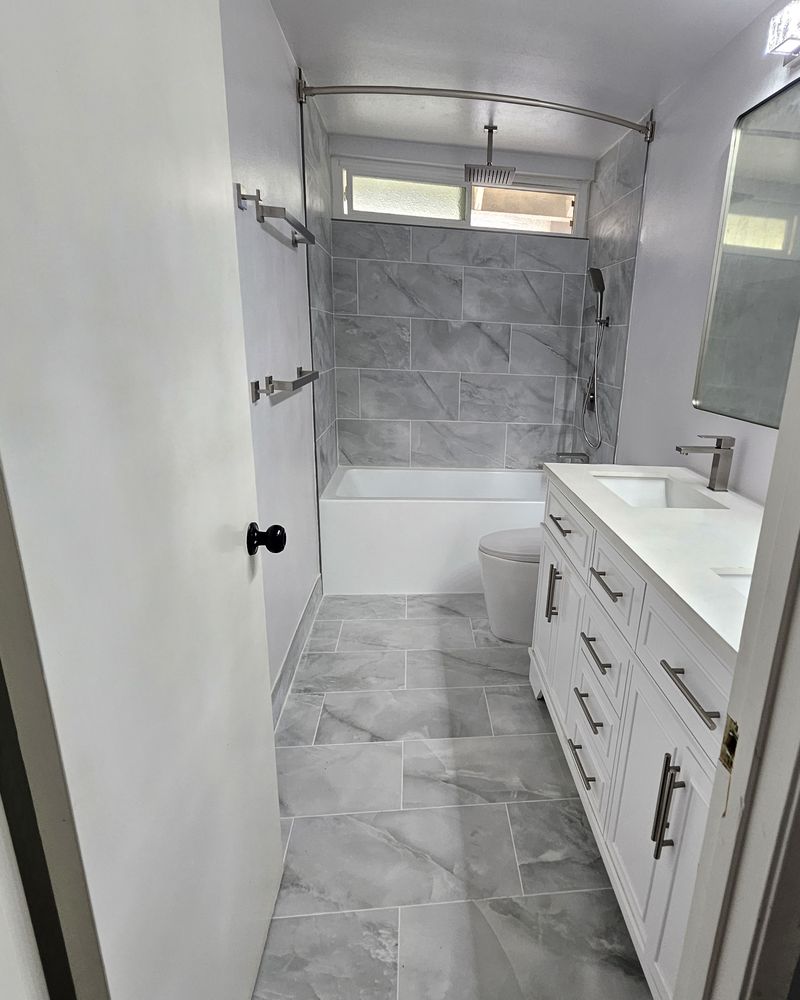 The same hall bathroom finished, with a tiled tub surround, gray marble look floor tile, and a white double vanity